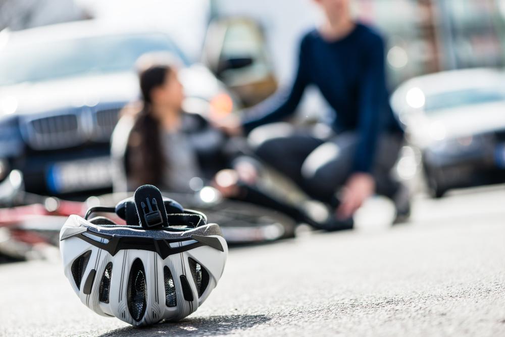 Close-up of a bicycle helmet on the road with someone in the background helping a woman who was hit by a car while bicycling.