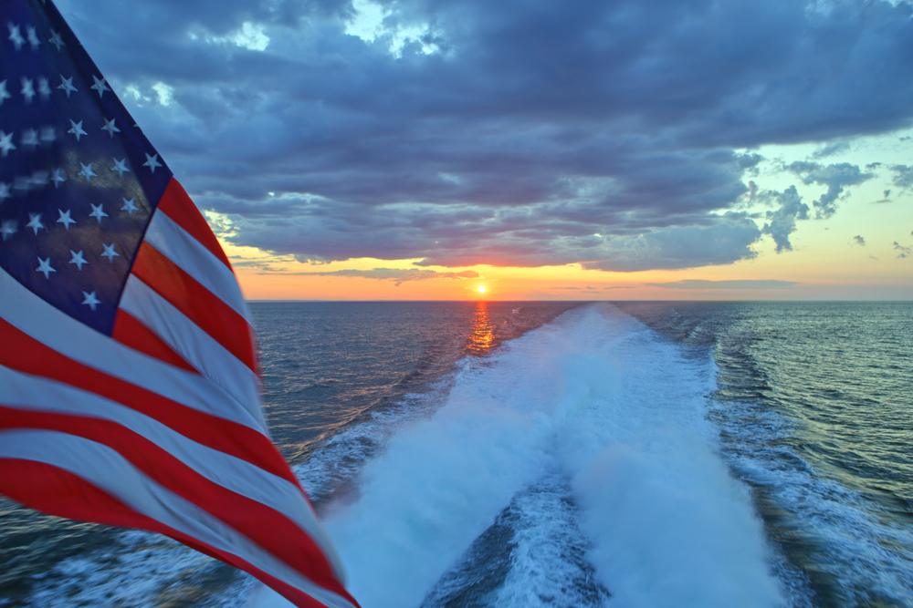 American flag waving on a boat, with the water and sunset in the background.
