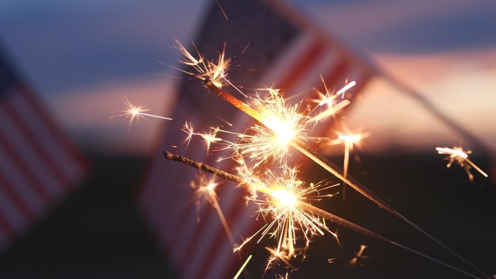 People holding lit sparklers in front of a US flag on the Fourth of July.